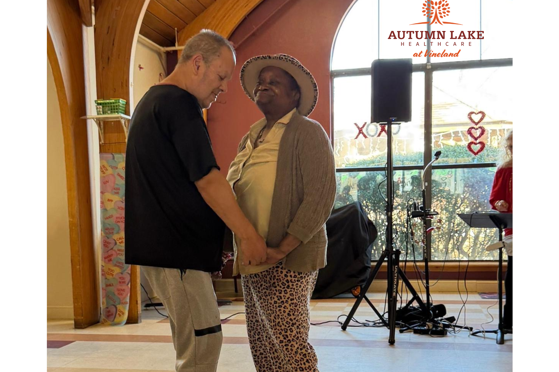Two residents hold hands and dance together in a brightly lit common room during a social event at an assisted living facility.